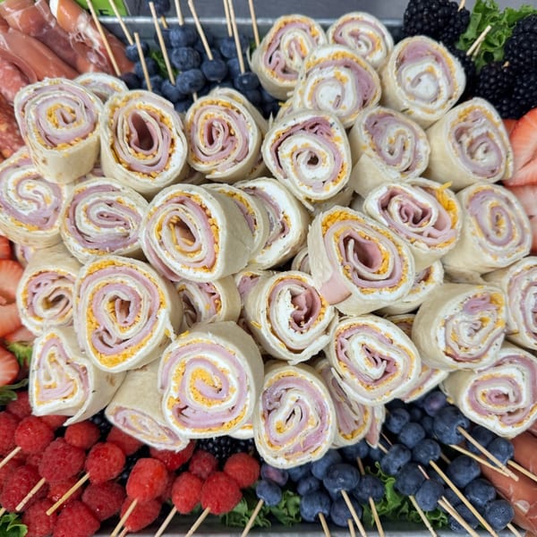 A platter filled with pinwheel wraps, strawberries, raspberries, blueberries, and charcuterie.
