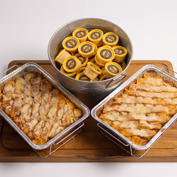 Two foil pans of peach cobbler and a bucket of Blue Bell ice cream cups on a wooden board.