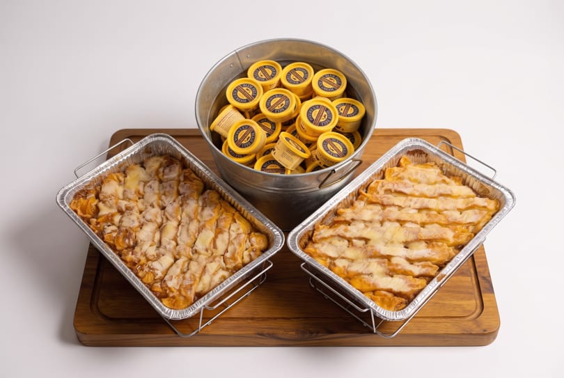 Two foil pans of peach cobbler and a bucket of Blue Bell ice cream cups on a wooden board.