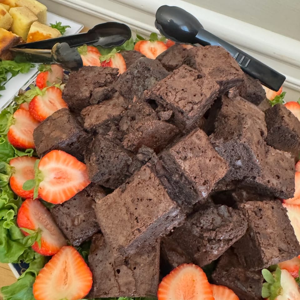 A platter of brownies and sliced strawberries on a bed of lettuce with serving tongs.