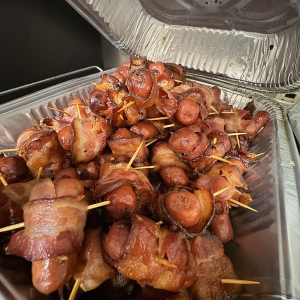 A foil pan filled with numerous bacon-wrapped hot dogs, each held by a toothpick.